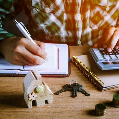 Selective focus wood home key and step up gold coin or currency on wooden table with document.Blur hand man calculate finance and writing on notebook at home office desk.business concept background.